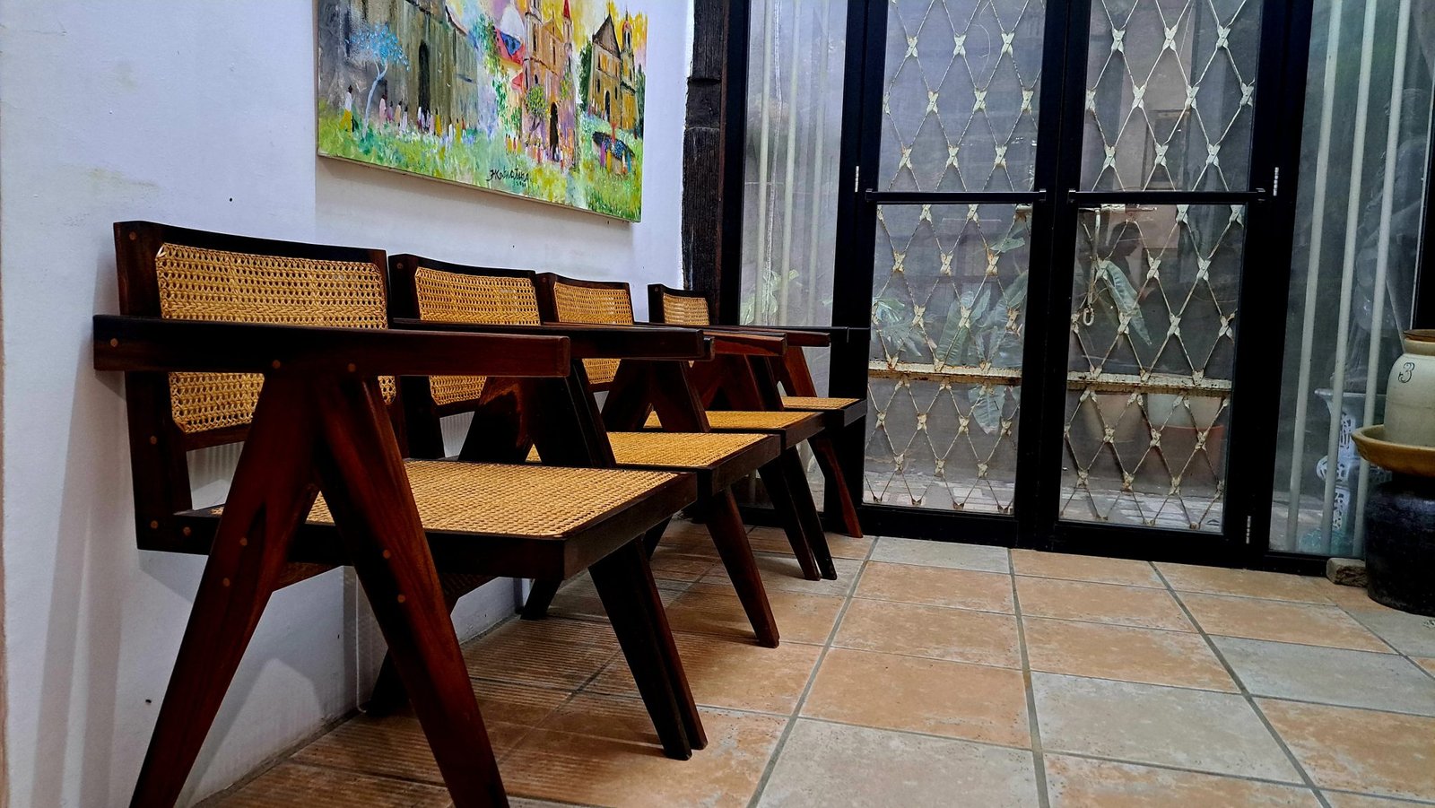A row of cane and wood chairs against a wall beside a window, with a painting hung above them.